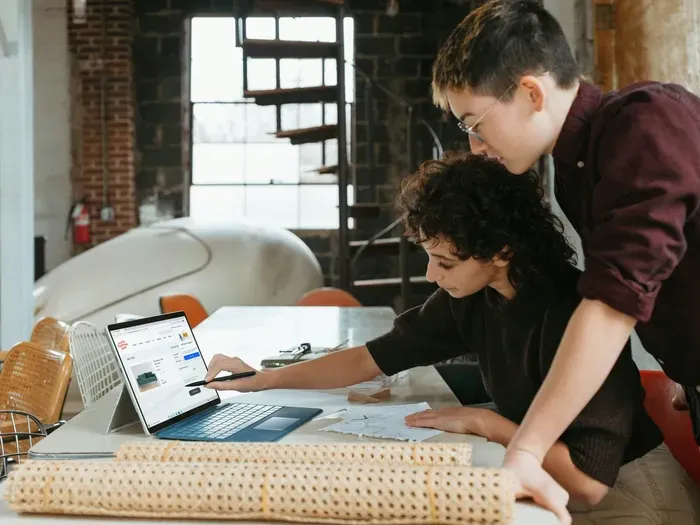 Two young professionals collaborating at a desk, reviewing blueprints and laptop for creative project